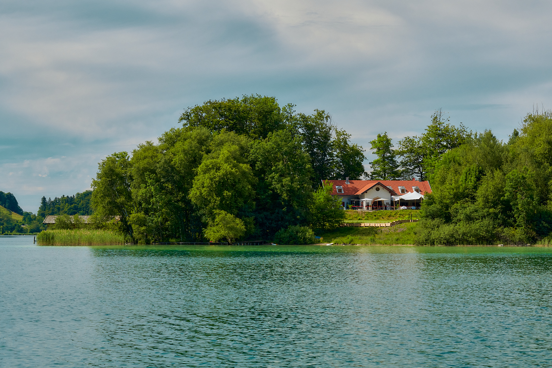 Die Insel vom Wasser aus: das Haus mit rotem Ziegeldach und Sonnenschirmen zwischen altem Baumbestand